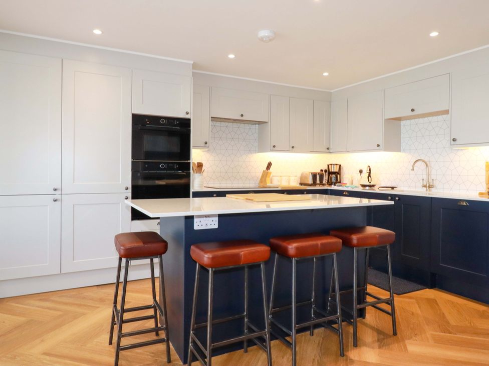 A kitchen with white and dark blue cabinets a white countertop an island with four brown stools and wooden flooring at Pink House in Mevagissey