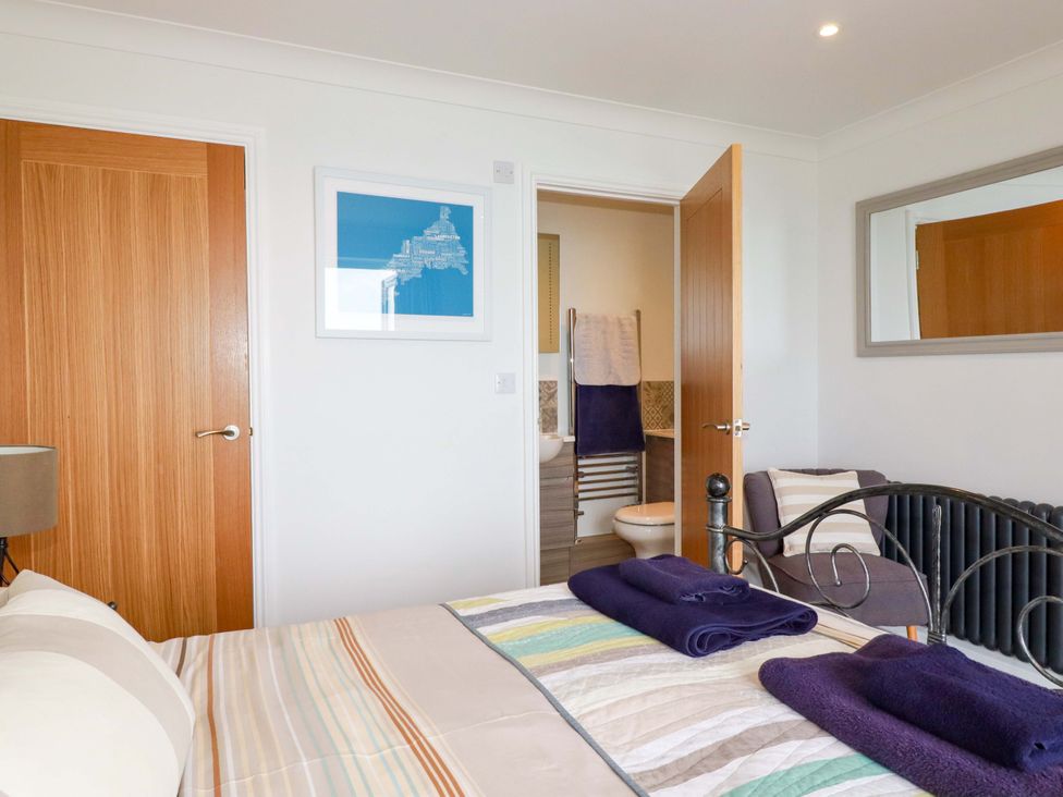 A bedroom with a bed, towels on the bed, two wooden doors, a chair with a cushion, a wall mirror and an en suite bathroom at Pink House Mevagissey