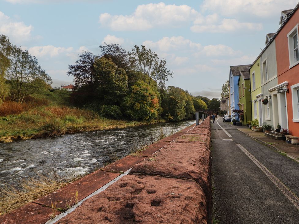 A riverside view with a walkway and colorful houses at Riverside Cottage Cockermouth