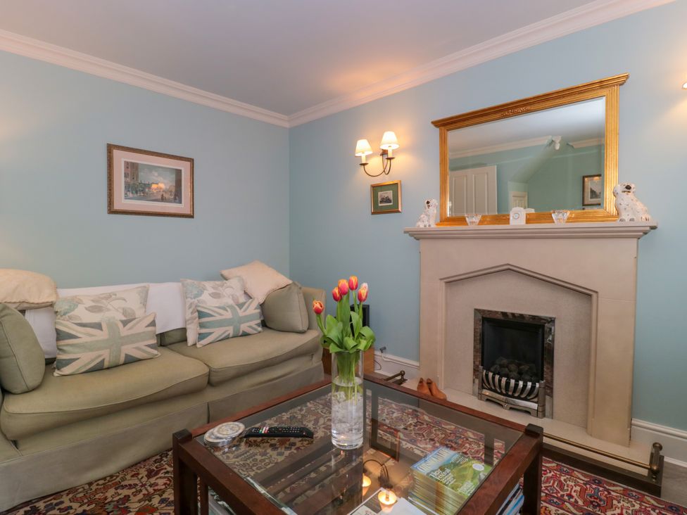 A living room with a green sofa, cushions, a glass coffee table with flowers, and a fireplace with a mirror above at Batemans Nailsworth