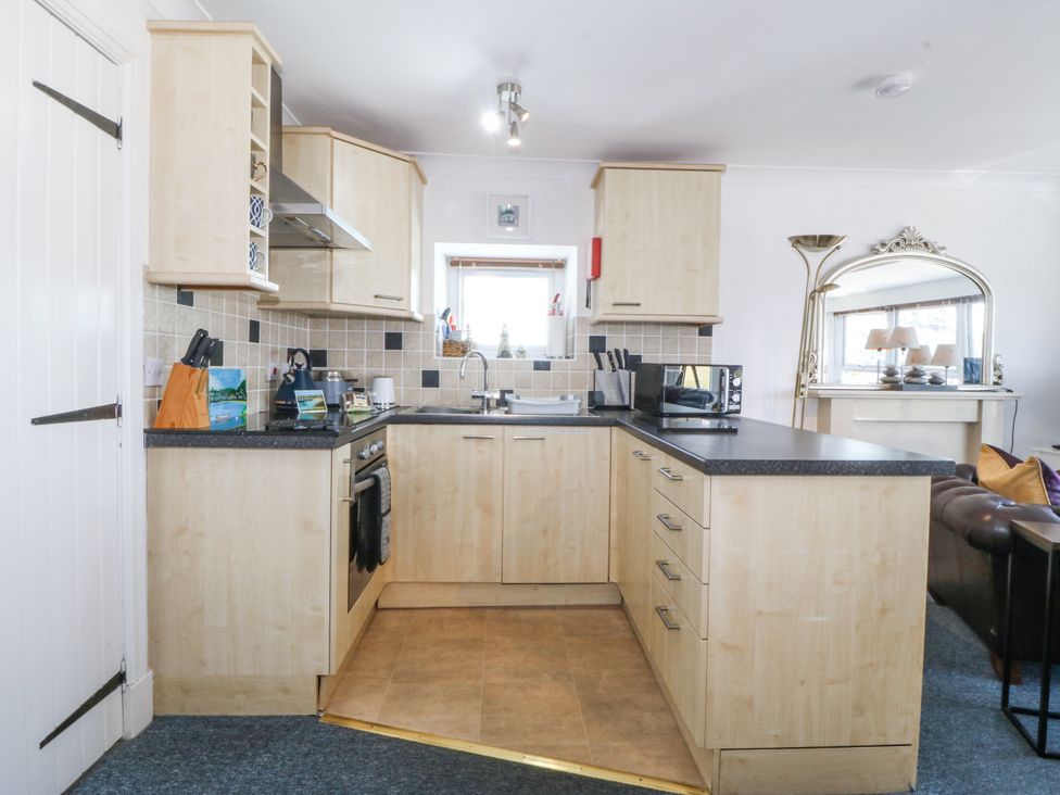 A kitchen with light wood cabinets, a black countertop, a microwave, a kettle, a sink below a window, and a tiled backsplash at Sea View apartment in Borth-Y-Gest
