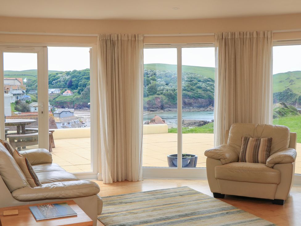 A living room with beige leather sofas and large windows overlooking a patio and green hills at 2 Sea Gardens in Hope Cove