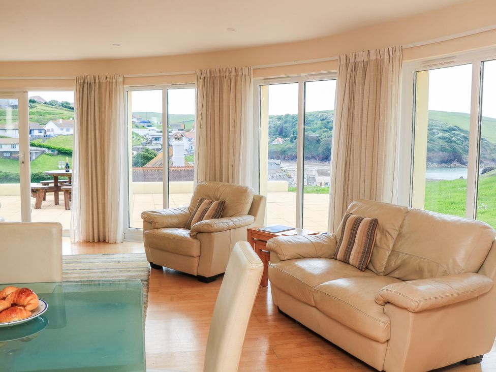 A living room with beige armchairs and sofa near large windows showing a grassy landscape at 2 Sea Gardens in Hope Cove