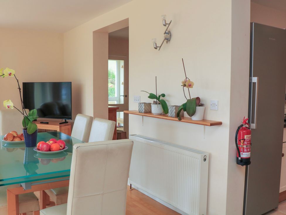 A dining area with a glass table and beige chairs next to a TV and a wall shelf with plants at 2 Sea Gardens in Hope Cove