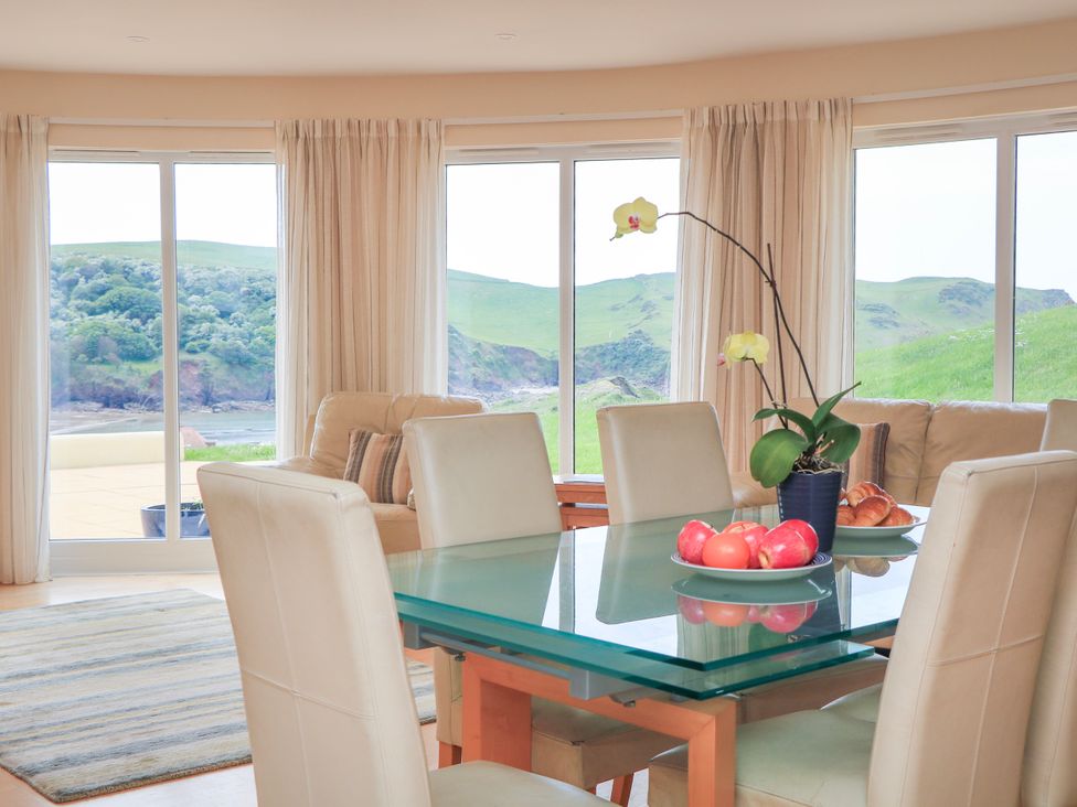 A dining area with a glass table and beige chairs with a flower pot and fruit plates near large windows showing green hills at 2 Sea Gardens Hope Cove