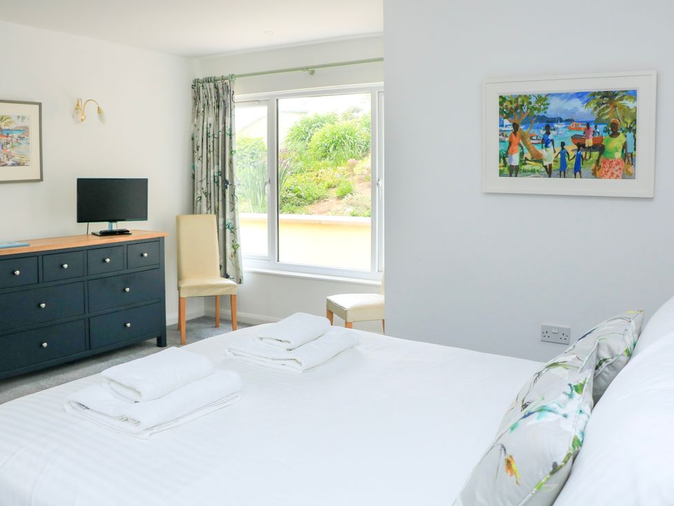 A bedroom with a bed with folded towels a window two chairs a dresser with a TV and paintings on the walls at 2 Sea Gardens in Hope Cove