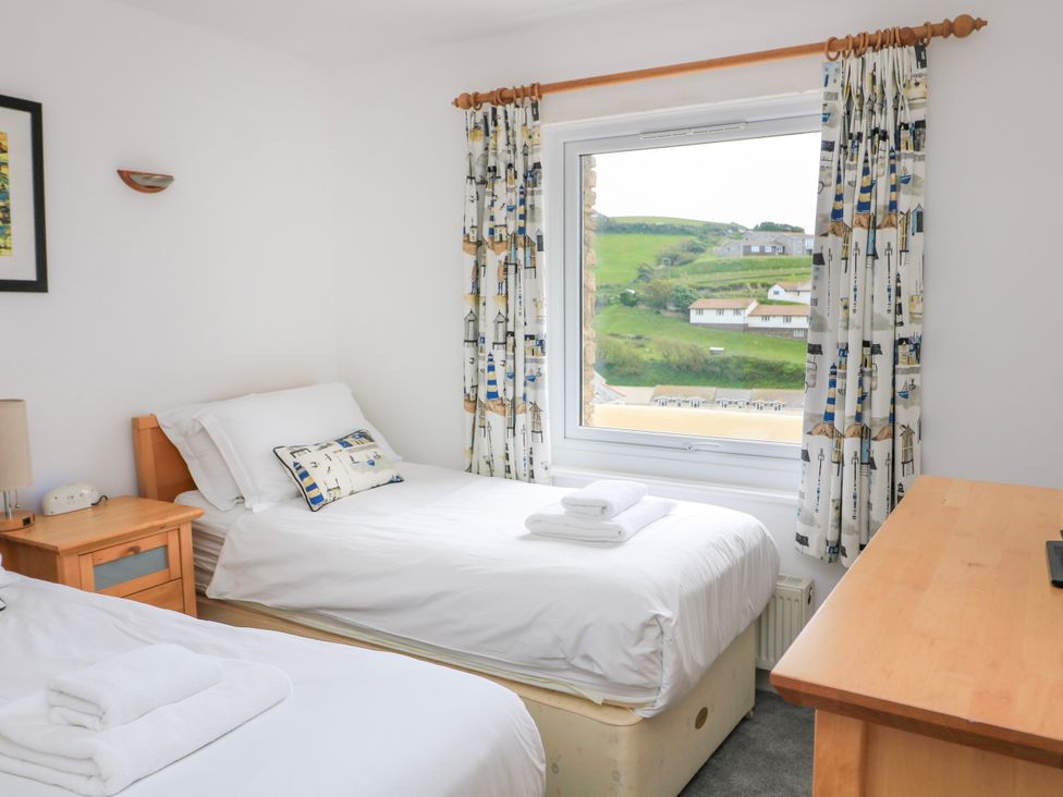 A bedroom with two single beds a wooden desk a window with curtains overlooking green hills at 2 Sea Gardens in Hope Cove