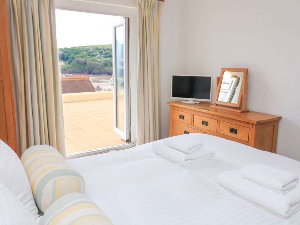 A bedroom with a bed, white linens, two bolsters, a wooden dresser with a mirror and TV, and an open door leading to a balcony at 2 Sea Gardens in Hope Cove
