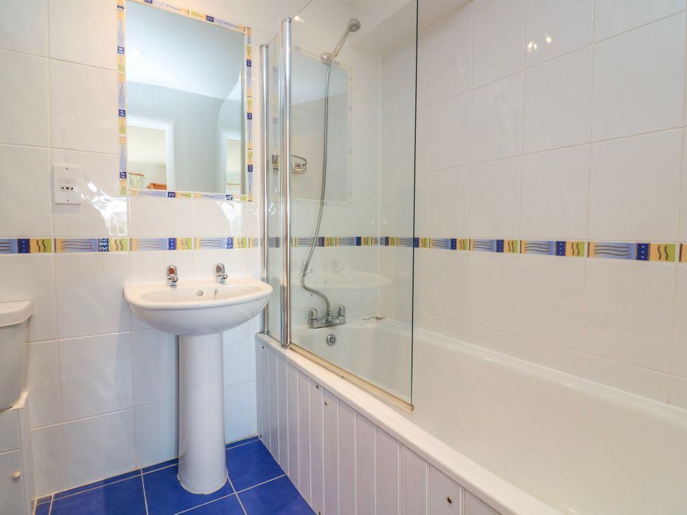A bathroom with a sink a mirror a bathtub with glass shower screen and white tiled walls with a colorful border at 2 Sea Gardens in Hope Cove
