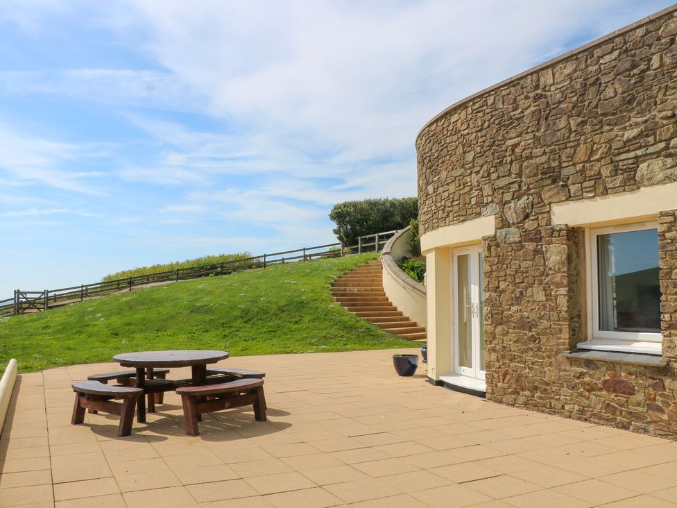 An outdoor patio area with a round wooden picnic table and curved stone building with steps leading up a grassy hill at 2 Sea Gardens in Hope Cove